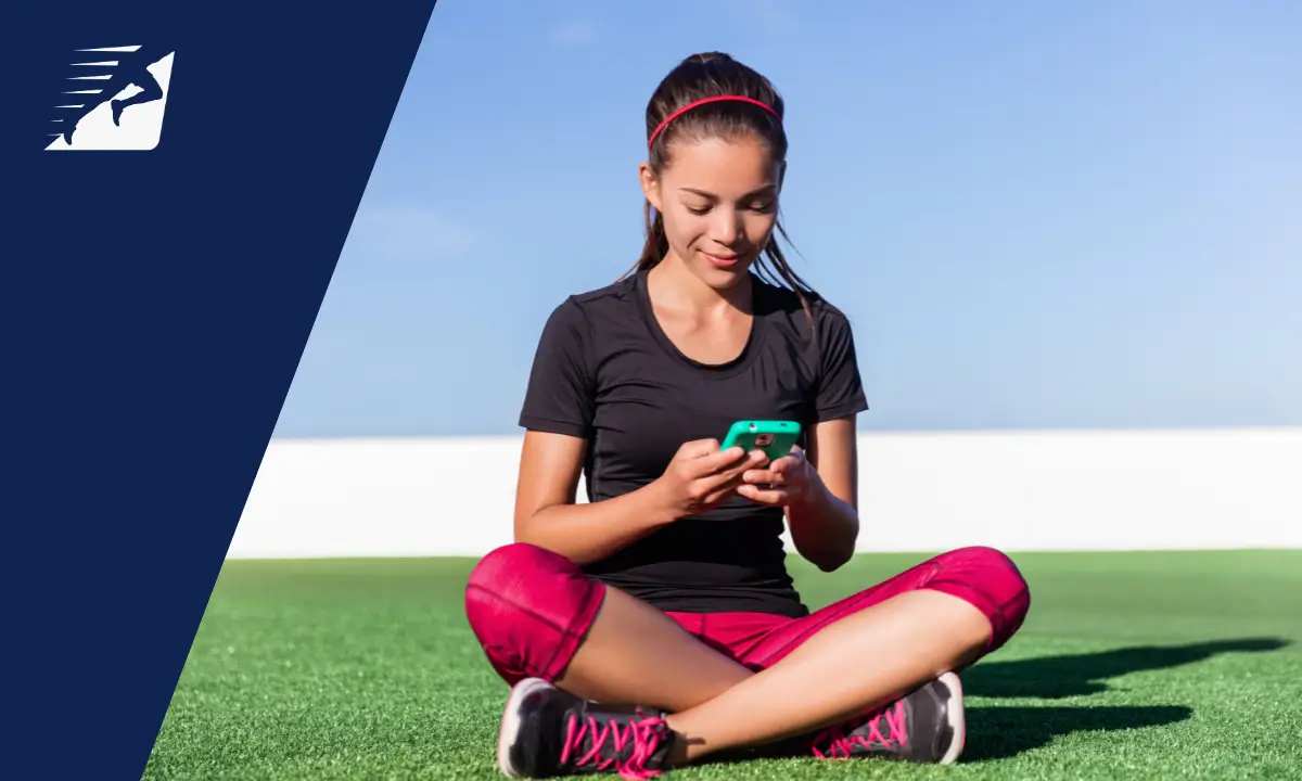 Eine sportlich bekleidete Frau sitzt auf einem Rasen und bedient ein Smartphone